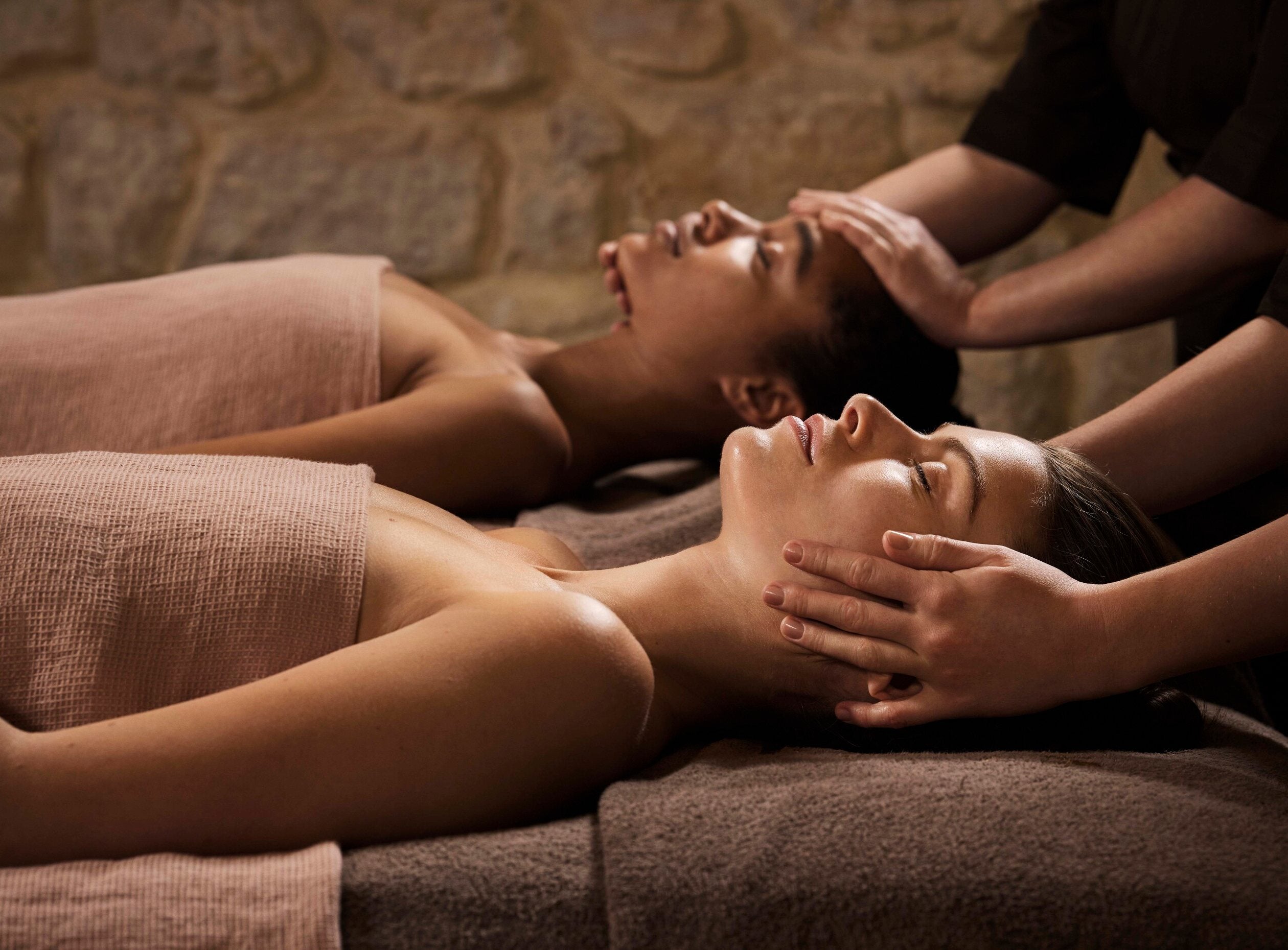 deux femmes avec une serviettes nouées autour de leur corps allongées sur le dos, sur deux tables de massage différentes. L'une est en train de se faire masser les cheveux tandis que l'autre se fait masser le visage. Elles ont les yeux fermés et semblent détendues