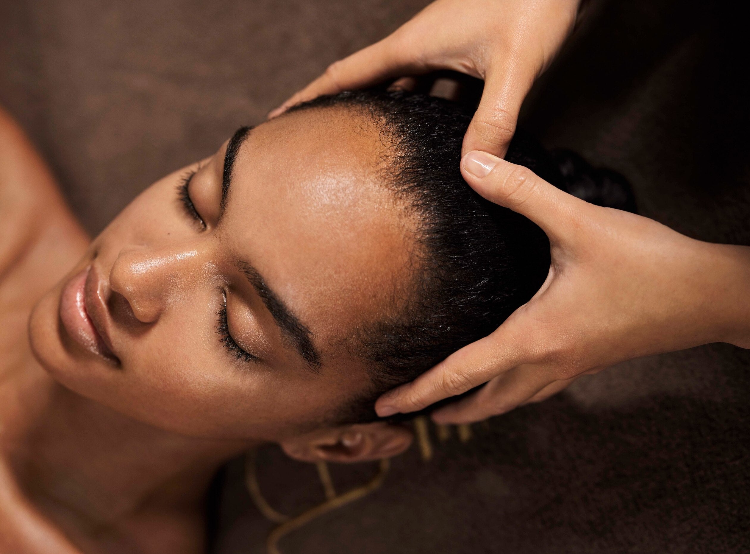 visage d'une femme en train de se faire masser le cuir chevelu. Elle ferme les yeux et sourit légèrement. Elle semble apaisée