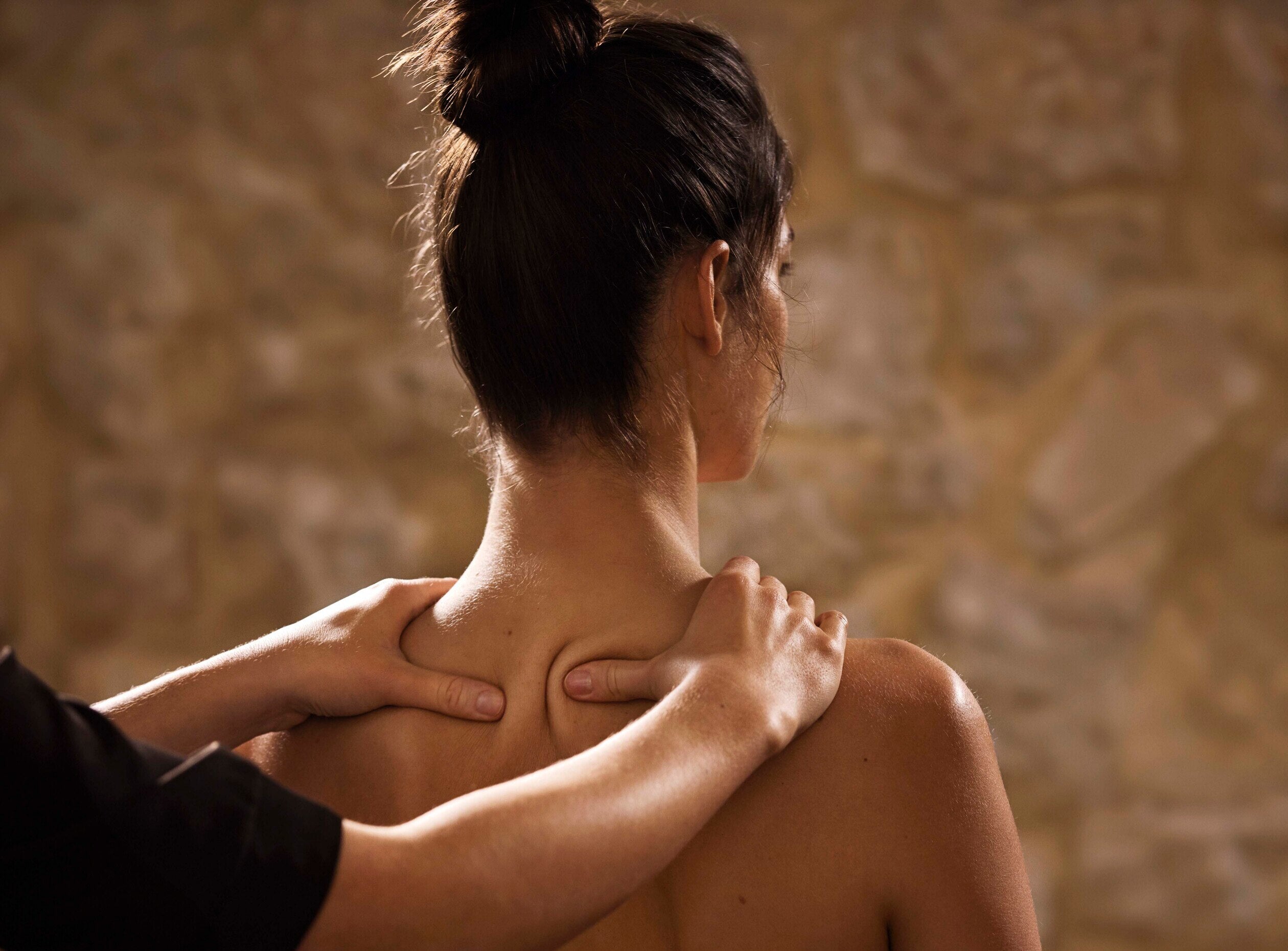 tête et épaules d'une femme de dos. ses cheveux sont relevés en chignon. Elle est en train de se faire masser les épaules (trapèzes) par une autre personne