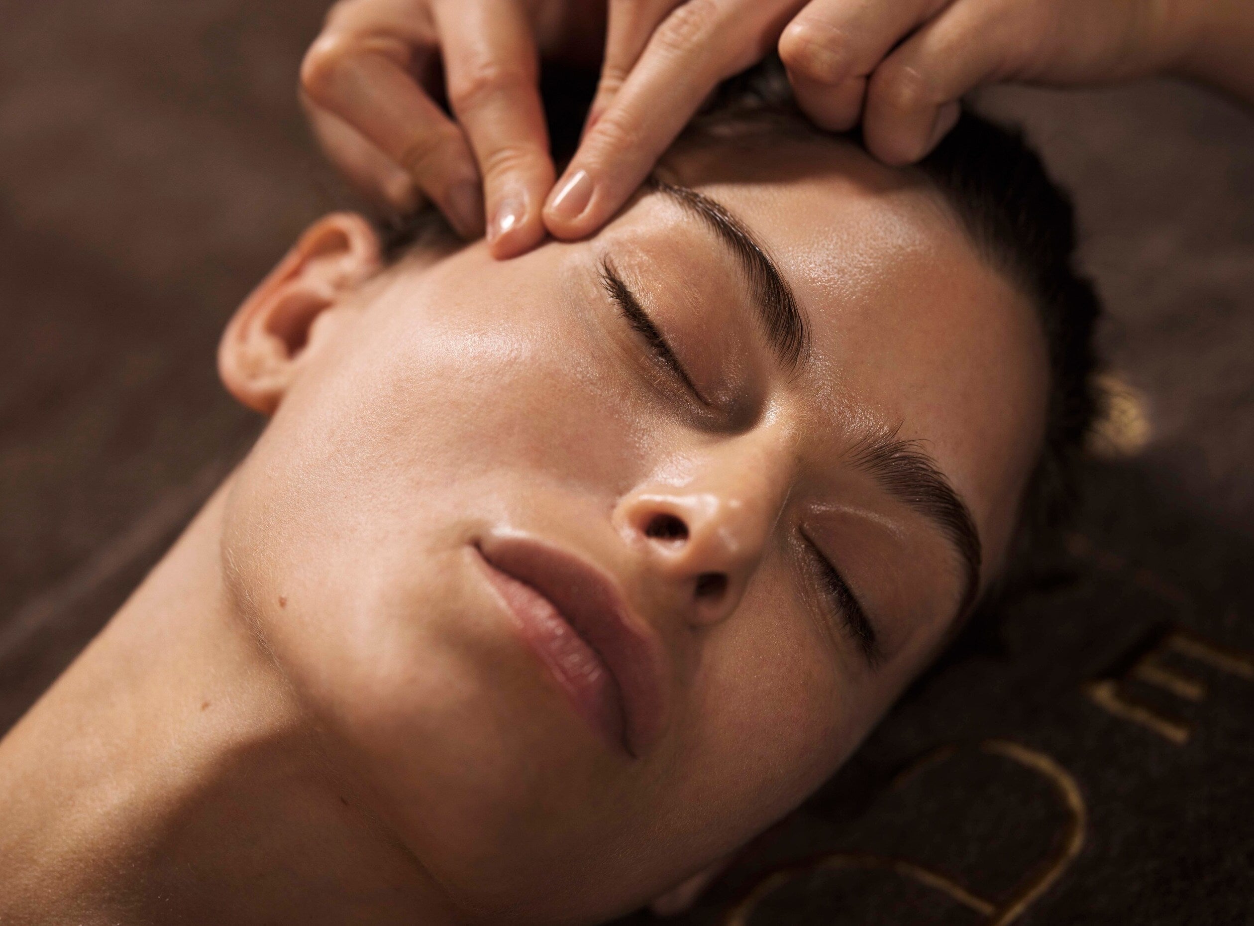 visage d'une femme reposant sur une serviette marrons. Elle ferme les yeux et semble détendue. deux doigts lui massent le contour des yeux. C'est une scène de massage de soin visage.