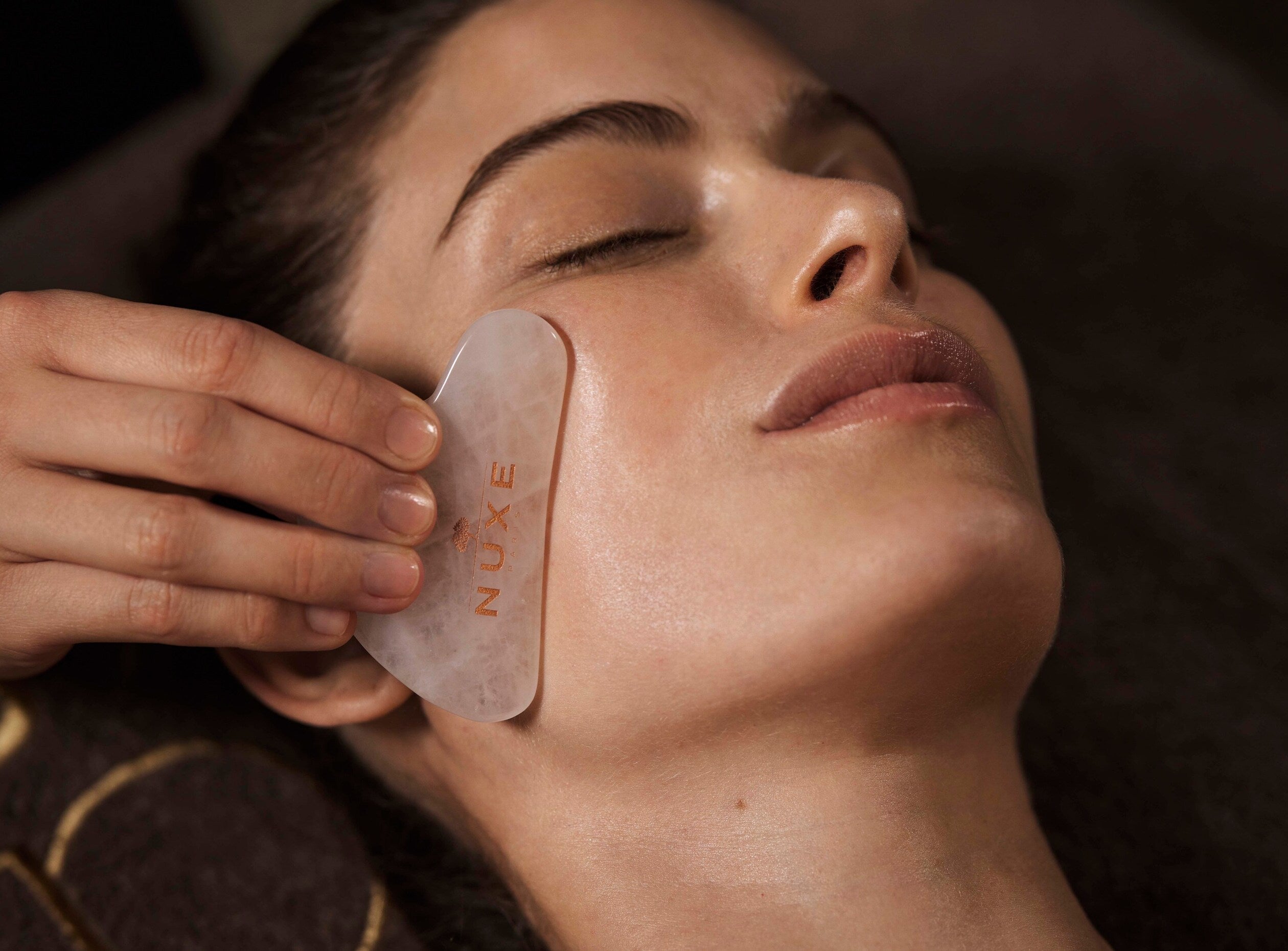visage d'une femme en train de se faire masser le côté de son visage grâce à une gua sha (outil de massage). Elle ferme les yeux. L'ambiance et tamisée et détendue, propre à un spa