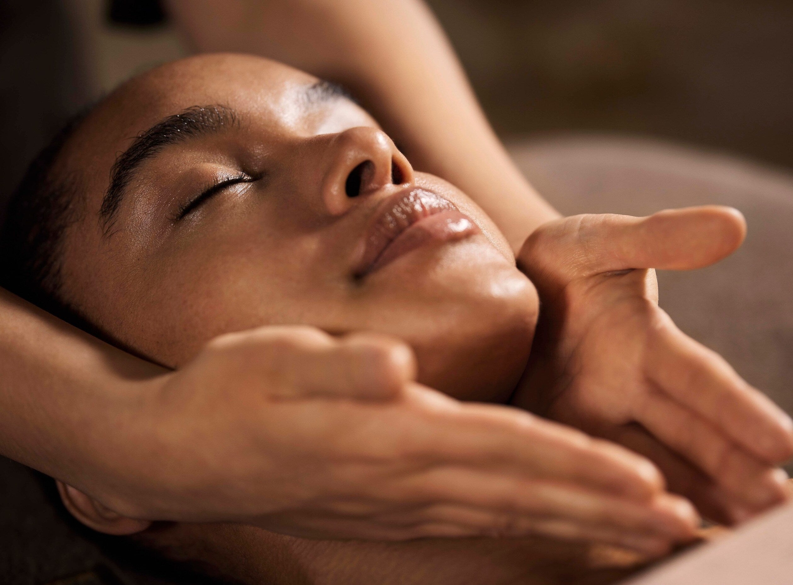 visage d'une femme en train de se faire masser l'ovale de son visage. Elle ferme les yeux et sourit légèrement. Ambiance tamisée et détendue, propre à un spa.