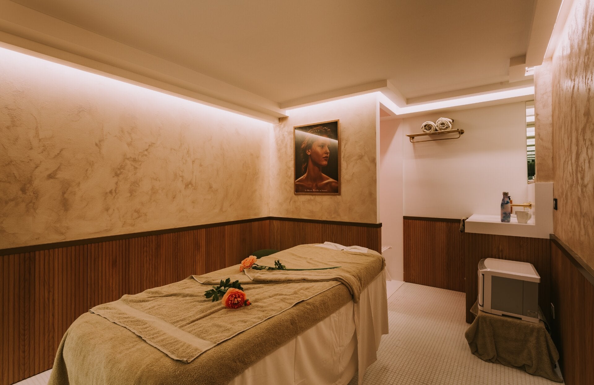 Une cabine de soin intime et élégante, baignée d’une lumière douce et chaleureuse. La table de massage, délicatement préparée avec des linges soyeux et quelques fleurs, invite à un moment de détente absolue, dans une atmosphère apaisante propice au lâcher‑prise.