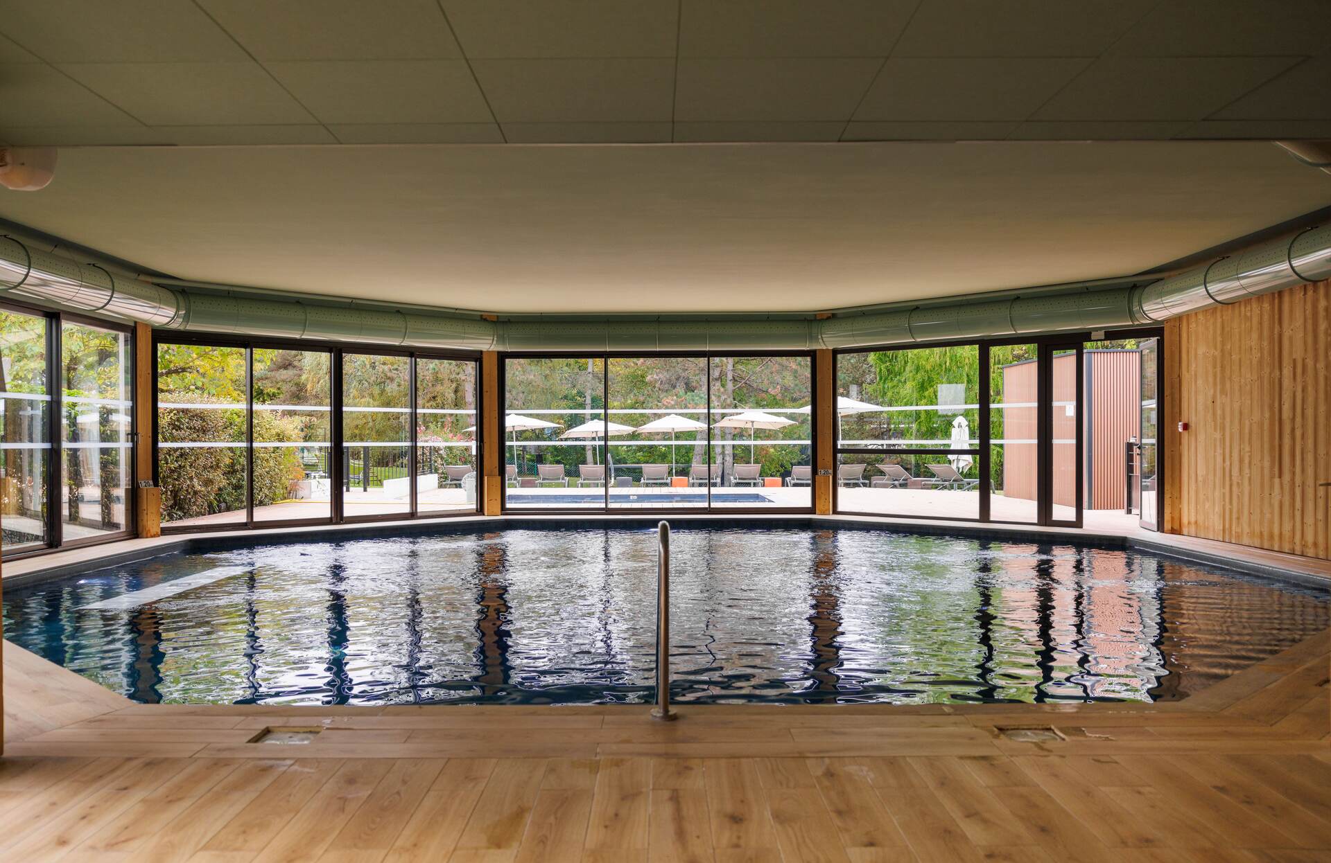 Un espace piscine intérieur élégant et contemporain, largement ouvert sur l’extérieur grâce à de grandes baies vitrées. Baigné de lumière naturelle et entouré de matériaux chaleureux comme le bois, ce lieu apaisant invite à la détente tout en offrant une vue agréable sur un environnement verdoyant.