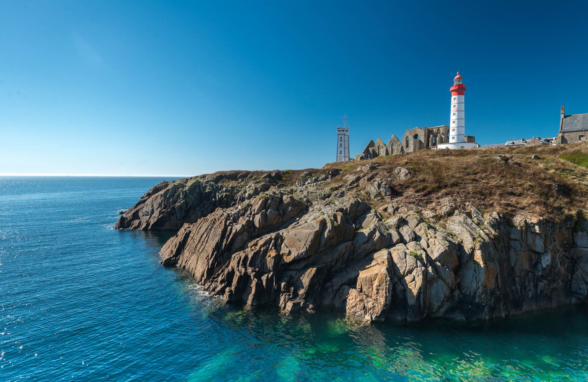 Un paysage côtier spectaculaire où un phare blanc et rouge se dresse fièrement au sommet de falaises rocheuses baignées par une mer aux reflets turquoise. Sous un ciel parfaitement dégagé, ce décor sauvage et préservé offre une atmosphère à la fois apaisante et majestueuse, invitant à l’évasion.
