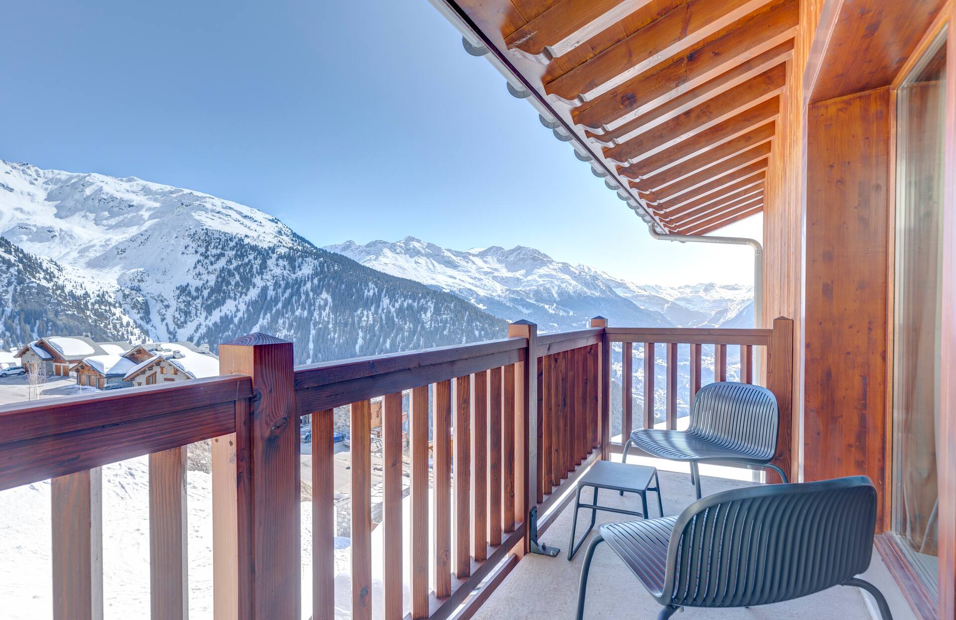Un balcon en bois chaleureux s’ouvre sur un panorama spectaculaire de montagnes enneigées, avec deux fauteuils design et une petite table invitant à la contemplation.
Les sommets majestueux, baignés de lumière sous un ciel pur, offrent une sensation d’évasion totale, idéale pour un séjour ressourçant au cœur d’un cadre alpin d’exception.