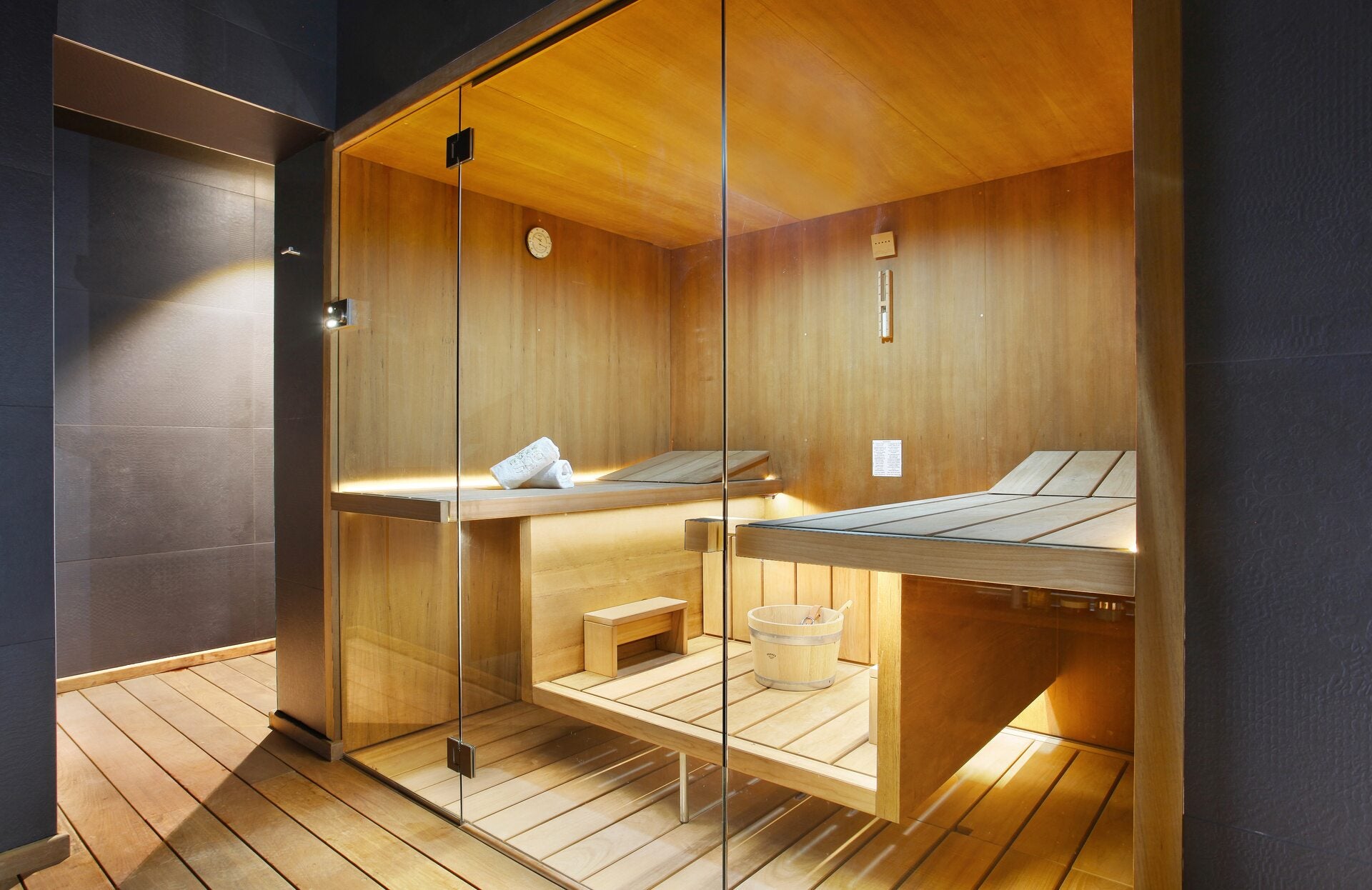 Un sauna moderne entièrement habillé de bois clair est visible derrière une paroi vitrée, avec des bancs disposés sur deux niveaux et un seau traditionnel posé au centre.
L’éclairage subtil intégré sous les assises met en valeur les textures chaleureuses du bois, créant une atmosphère apaisante et propice à un moment de relaxation intense.
