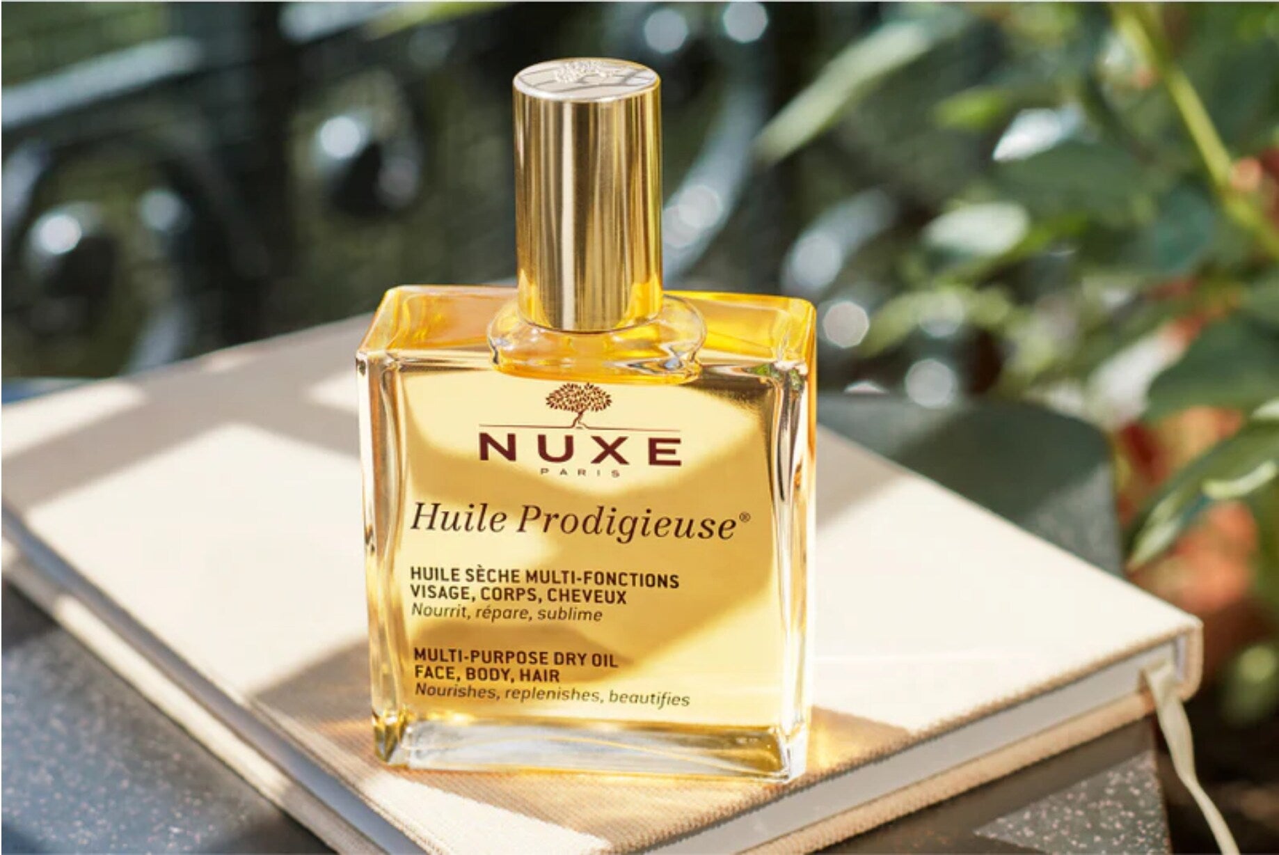 Un flacon carré en verre rempli d’une huile dorée est posé sur un carnet clair, avec le texte « NUXE Huile Prodigieuse » visible sur l’étiquette. Le bouchon doré brillant reflète la lumière du soleil.
L’arrière-plan est flou, composé de feuilles vertes et de lumière naturelle, créant une ambiance extérieure douce et lumineuse. Les reflets sur le verre et le liquide mettent en valeur l’aspect chaleureux et naturel du produit.
