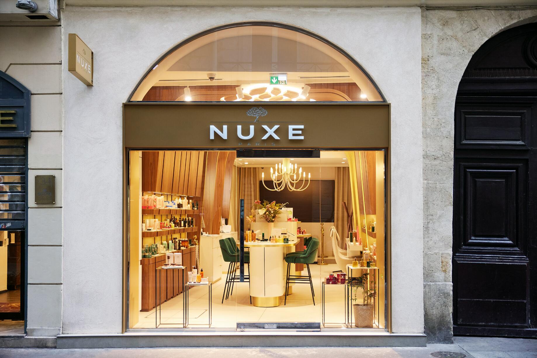 Une vitrine de boutique élégante est encadrée par une grande arche en verre, avec le nom « NUXE » affiché en lettres blanches au-dessus de l’entrée. L’intérieur est très lumineux, laissant apparaître un espace organisé et chaleureux.
À l’intérieur, des étagères en bois présentent de nombreux produits, tandis qu’un comptoir central entouré de chaises vertes attire le regard. Un lustre suspendu diffuse une lumière douce, renforçant l’ambiance raffinée et accueillante de la boutique.