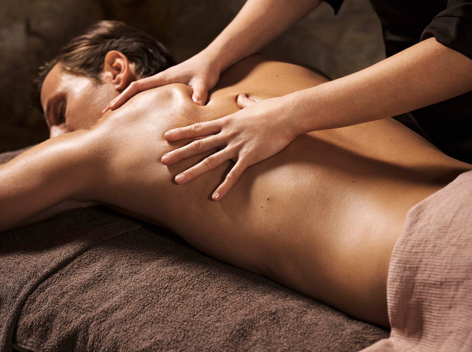 Un homme brun est allongé de profil sur le ventre, les yeux fermés dans une attitude de totale relaxation. Deux mains expertes massent fermement le haut de son dos nu et l'omoplate, sous une lumière douce et tamisée qui accentue l'ambiance apaisante du spa.