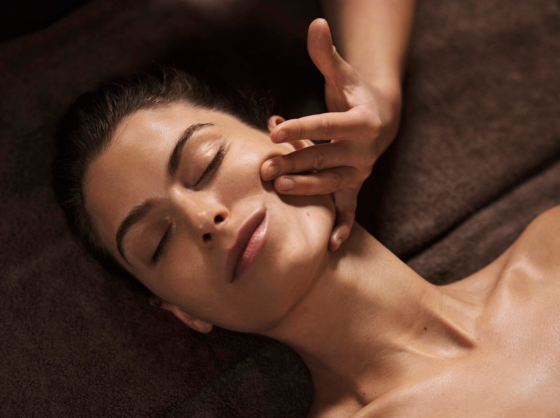Une personne est allongée sur une surface brune et douce, avec le haut du buste visible et la peau légèrement brillante. Une main effectue un massage du visage, en appuyant délicatement sur la joue avec les doigts.
La lumière chaude met en valeur les contours du visage et du cou, ainsi que la texture lisse de la peau. L’arrière-plan reste flou, renforçant l’ambiance calme et relaxante d’un soin en spa.