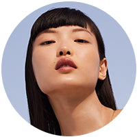 visage d'une femme sur fond ciel. Elle a la bouche légèrement ouverte. Elle est brune avec une frange