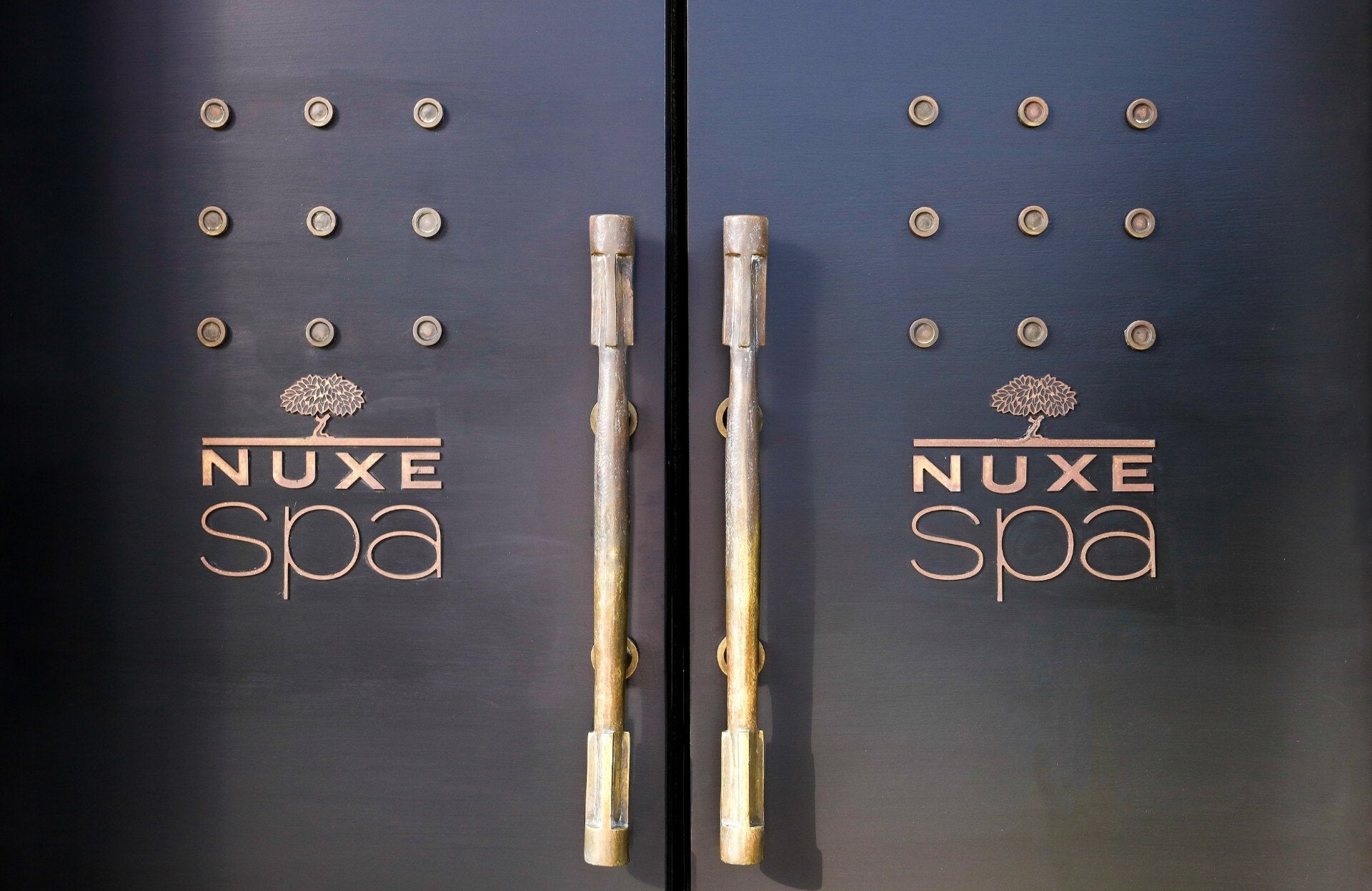 Deux grandes portes sombres occupent toute l’image, avec des poignées verticales en métal au centre. Chaque porte est décorée de petits reliefs circulaires disposés en grille, donnant un aspect texturé et élégant.
Le logo « NUXE spa » est inscrit en lettres dorées sur les deux battants, avec un arbre stylisé au-dessus du mot. L’ensemble est éclairé doucement, mettant en valeur le contraste entre le noir mat des portes et les reflets dorés des inscriptions et des poignées.