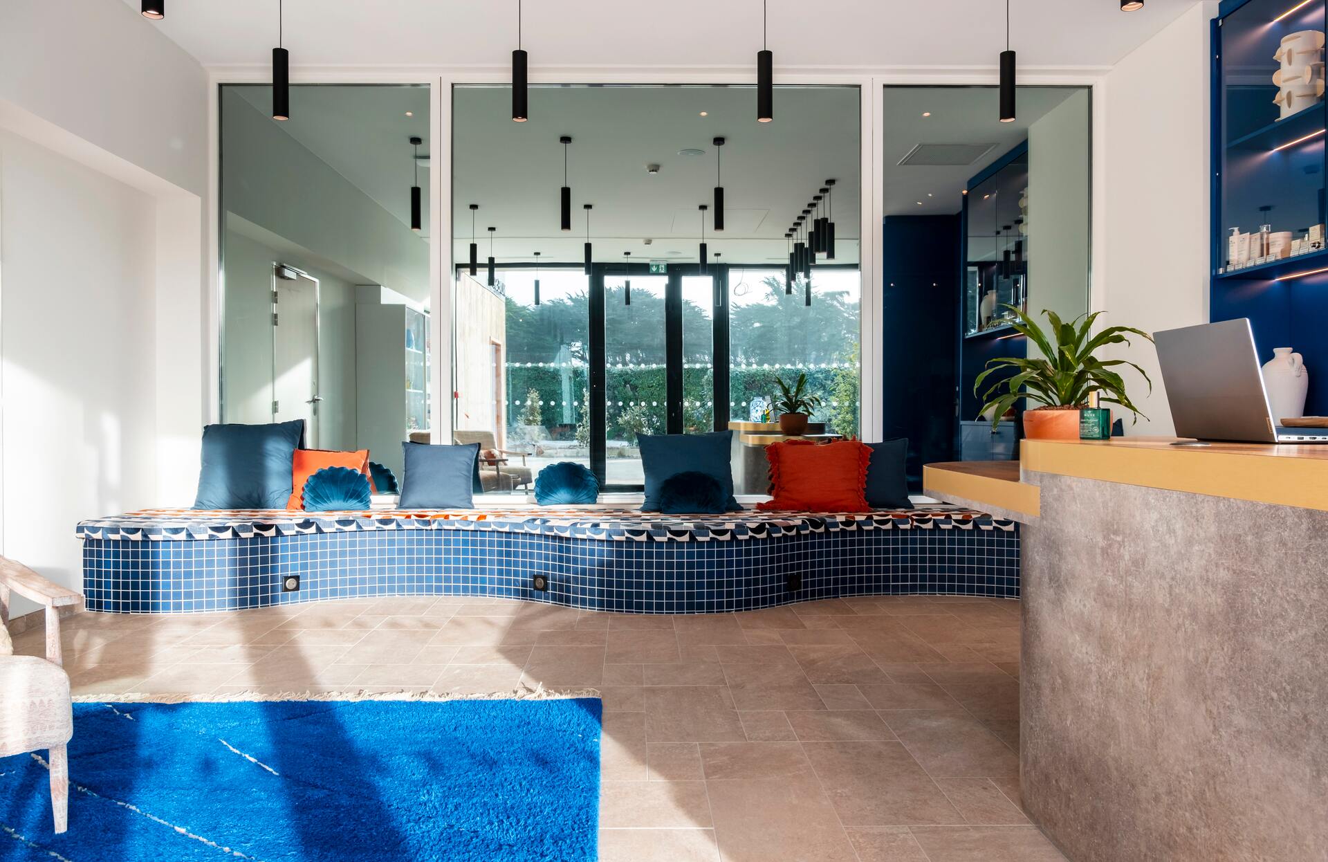 Un espace d’accueil contemporain et lumineux s’organise autour d’une banquette arrondie recouverte de coussins colorés, face à une large baie vitrée ouverte sur l’extérieur.
Les lignes épurées, les suspensions noires et le mélange de bois, de carrelage et de touches végétales créent une atmosphère élégante et immersive, idéale pour une expérience bien‑être moderne et apaisante.