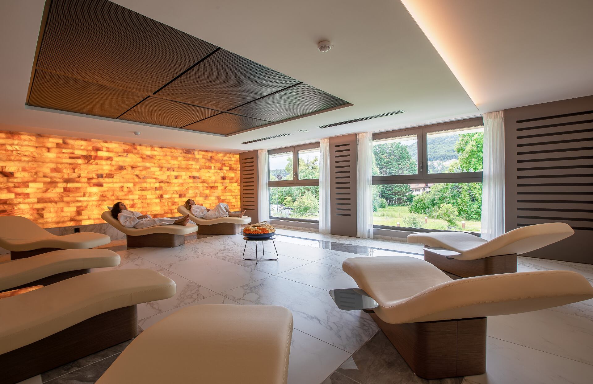 Un espace de relaxation baigné de lumière naturelle s’organise autour de larges fauteuils ergonomiques orientés vers de grandes baies vitrées ouvertes sur un paysage verdoyant.
Un mur rétroéclairé aux tons chauds diffuse une lumière enveloppante, créant une atmosphère apaisante et luxueuse, idéale pour un moment de détente absolue.