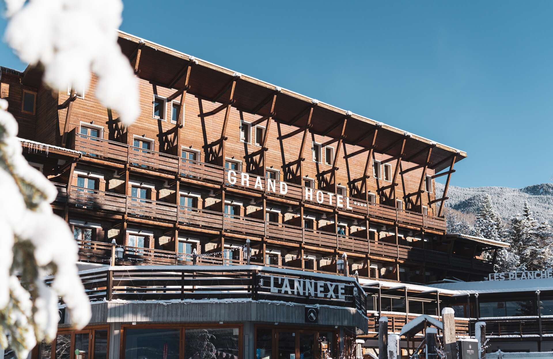 Un majestueux hôtel de style chalet, entièrement habillé de bois, se dresse au cœur d’un paysage enneigé sous un ciel bleu éclatant. La façade, rythmée par de nombreux balcons, affiche fièrement l’inscription “Grand Hôtel”, tandis qu’au premier plan, une annexe complète cet ensemble chaleureux et typiquement montagnard.