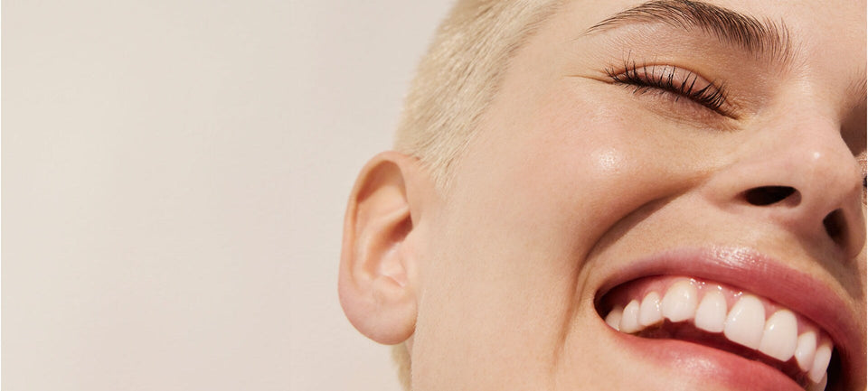 visage d'une femme sur fond beige. Elle est blonde, cheveux très courts, ses yeux sont fermés et elle affiche un grand sourire.