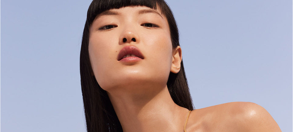 visage d'une femme sur fond bleu ciel. Elle a les cheveux noirs, la bouche légèrement ouvert. Sa peau est parfait et lumineuse