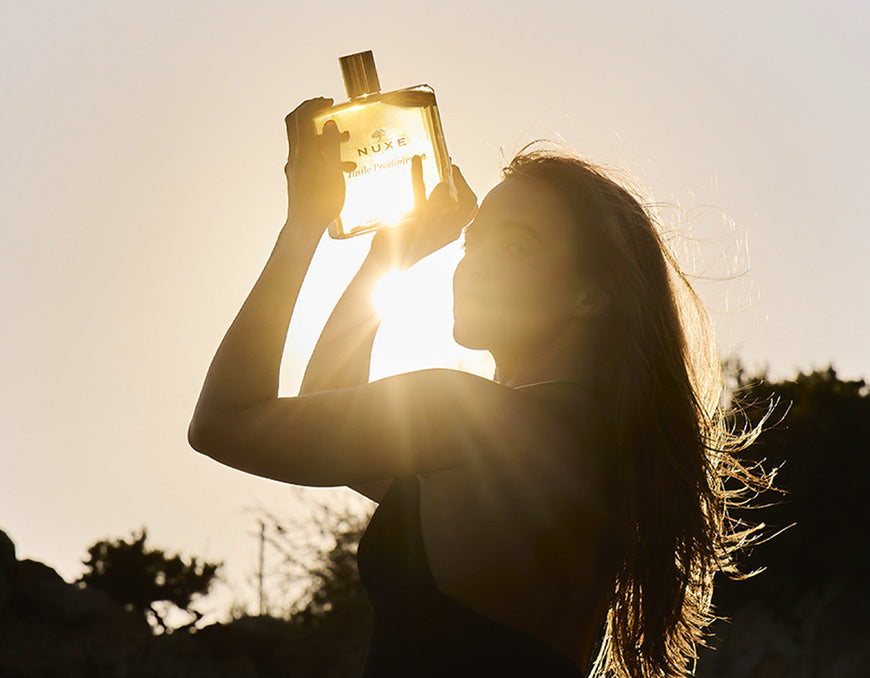 Femme en contre-jour au coucher du soleil, cheveux au vent, tenant à bout de bras un flacon d'Huile Prodigieuse NUXE traversé par la lumière dorée