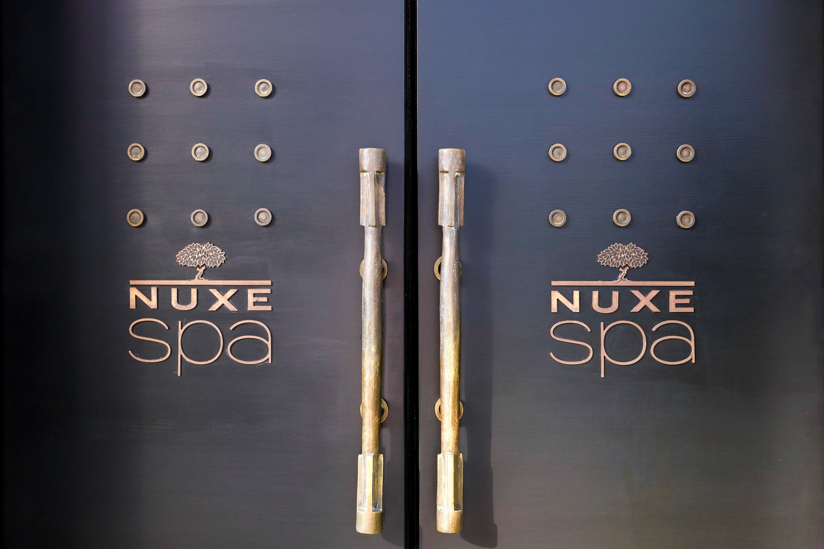 Deux grandes portes sombres occupent toute l’image, avec des poignées verticales en métal au centre. Chaque porte est décorée de petits reliefs circulaires disposés en grille, donnant un aspect texturé et élégant.
Le logo « NUXE spa » est inscrit en lettres dorées sur les deux battants, avec un arbre stylisé au-dessus du mot. L’ensemble est éclairé doucement, mettant en valeur le contraste entre le noir mat des portes et les reflets dorés des inscriptions et des poignées.