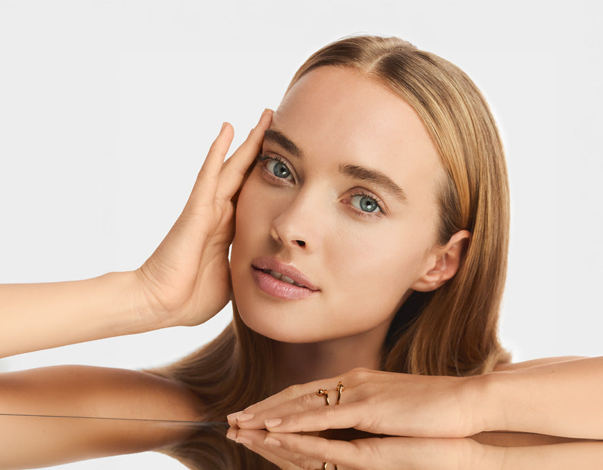 Portrait d'une femme aux cheveux blonds lisses et aux yeux clairs, une main sur la tempe, le menton posé sur ses mains au-dessus d'une surface miroir