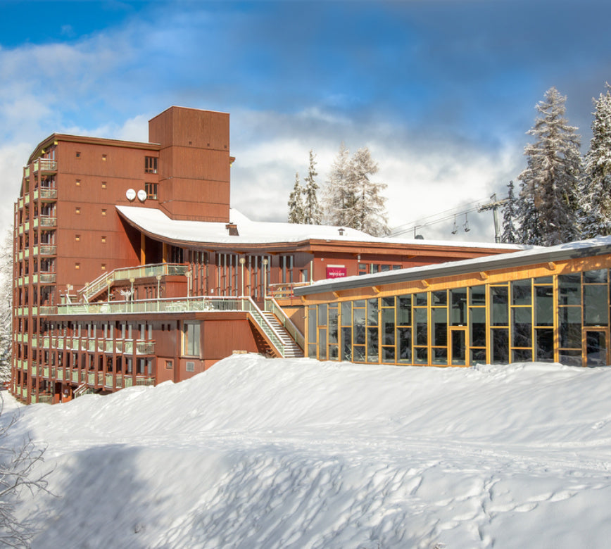 Vue lumineuse de la façade du spa Altezza Les Arcs, à l’architecture moderne et chaleureuse en bois, entourée d’une neige abondante et pure, sous un ciel bleu clair et apaisant en montagne. 