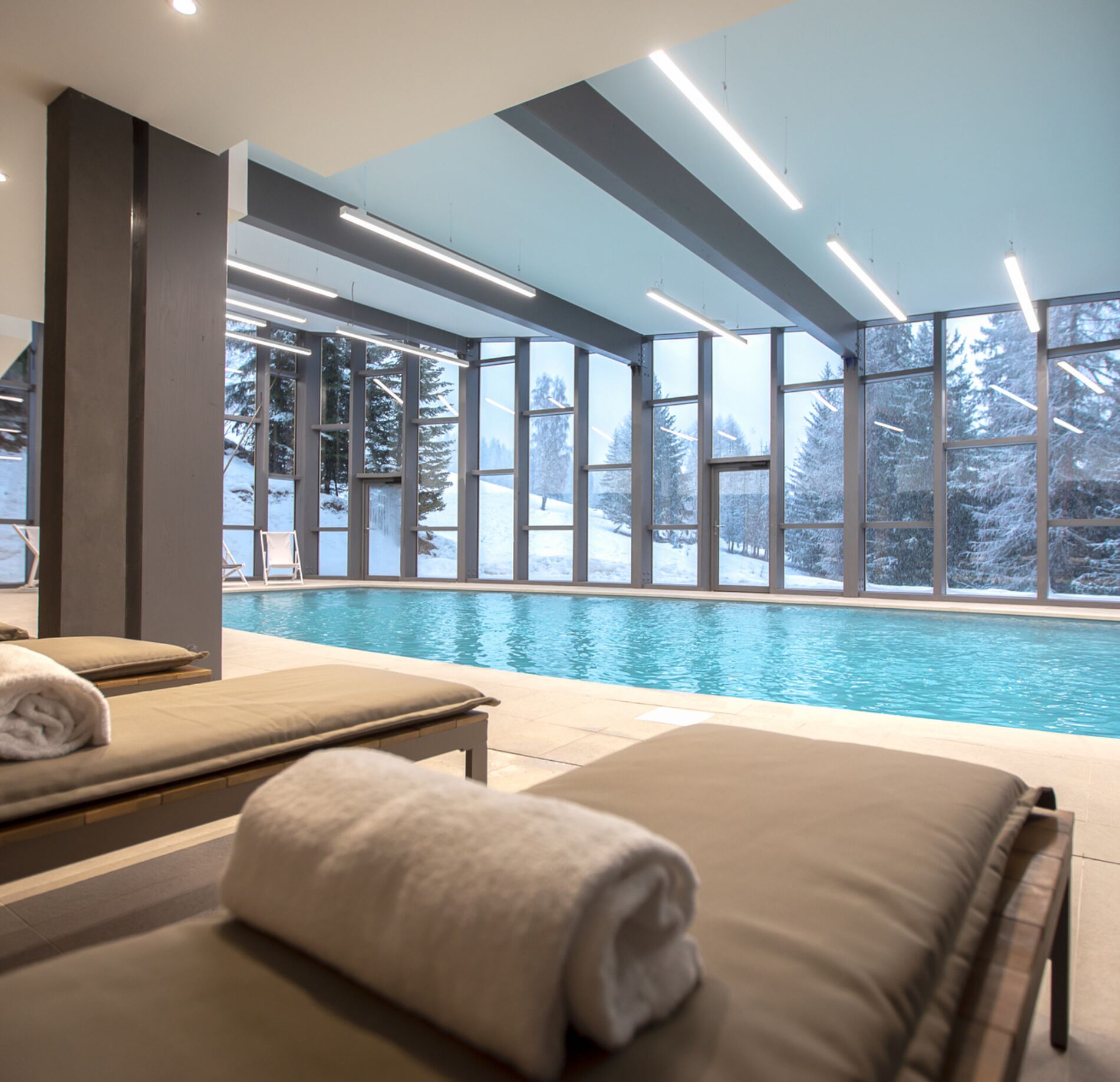 Piscine intérieure lumineuse du spa Altezza Les Arcs, bordée de larges baies vitrées offrant une vue apaisante sur un paysage enneigé et des sapins majestueux