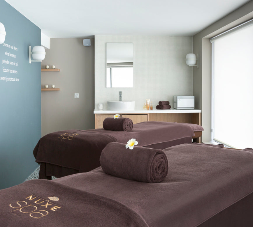 Salle de massage élégante du spa NUXE à l’Hôtel L’Araucaria, avec deux tables de soin confortables, mobilier en bois clair, espace lavabo minimaliste et grande baie vitrée diffusant une lumière naturelle, créant une ambiance sereine et haut de gamme. 