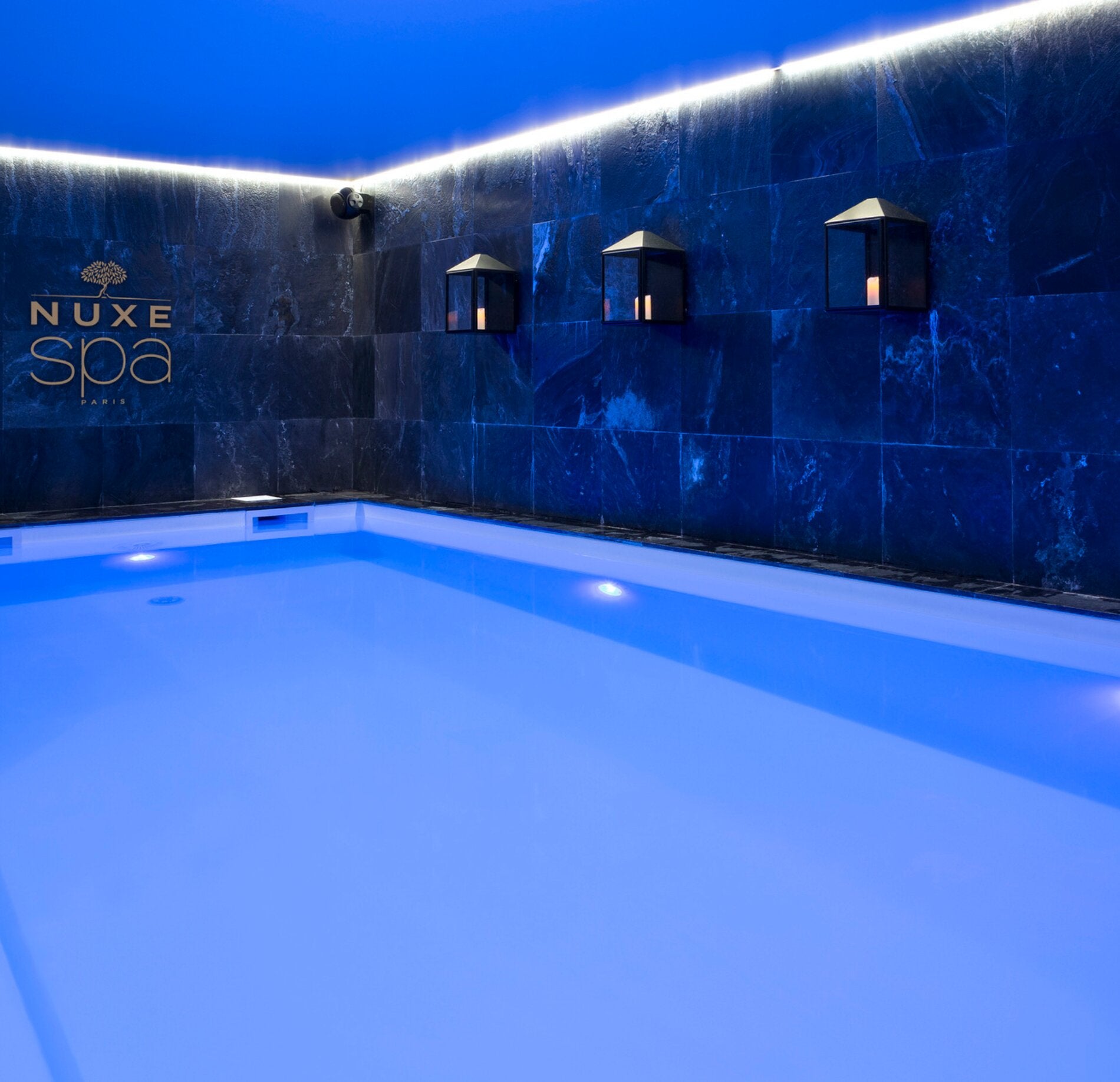 Piscine intérieure élégante du spa NUXE à l’Hôtel L’Araucaria, avec eau lumineuse aux reflets bleutés, mur en pierre sombre décoré de lanternes chaleureuses et éclairage indirect créant une atmosphère calme, intime et raffinée.