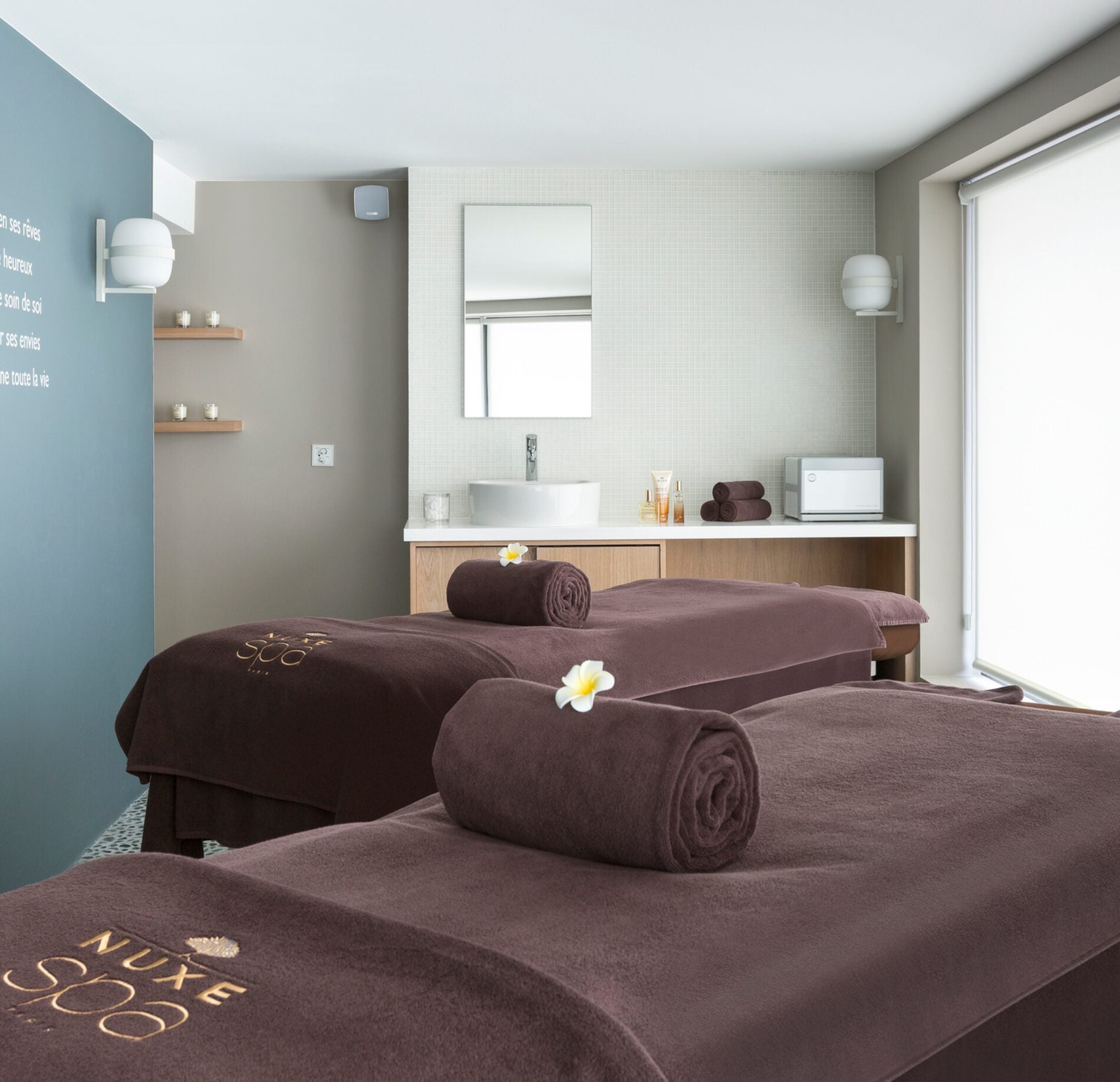 Cabine de soin lumineuse et contemporaine du spa NUXE de l’Hôtel L’Araucaria, avec tables de massage habillées de linge brun élégant, rouleaux de serviettes soignés et touches florales, dans une atmosphère douce, épurée et apaisante. 