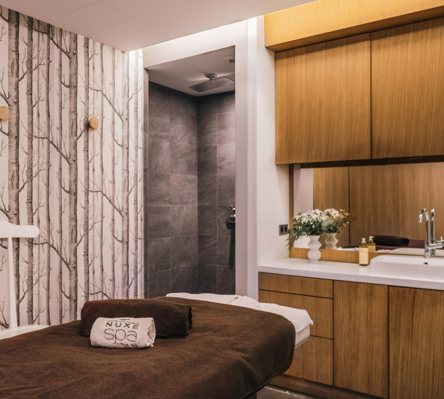 Salle de massage cosy du spa NUXE à l’Hôtel Balthazar, mêlant matériaux naturels et tons apaisants, avec table de soin confortable, mobilier en bois, espace de préparation équipé et papier peint décoratif, créant une ambiance intime et haut de gamme propice à la détente.