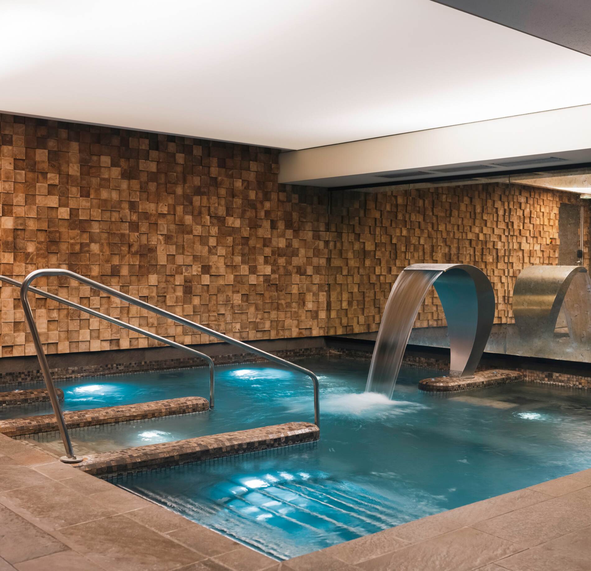 Bassin intérieur de balnéothérapie du spa NUXE à l’Hôtel Balthazar, avec eau turquoise apaisante, fontaines en cascade élégantes en inox et mur en bois texturé créant une atmosphère chaleureuse, raffinée et profondément relaxante. 