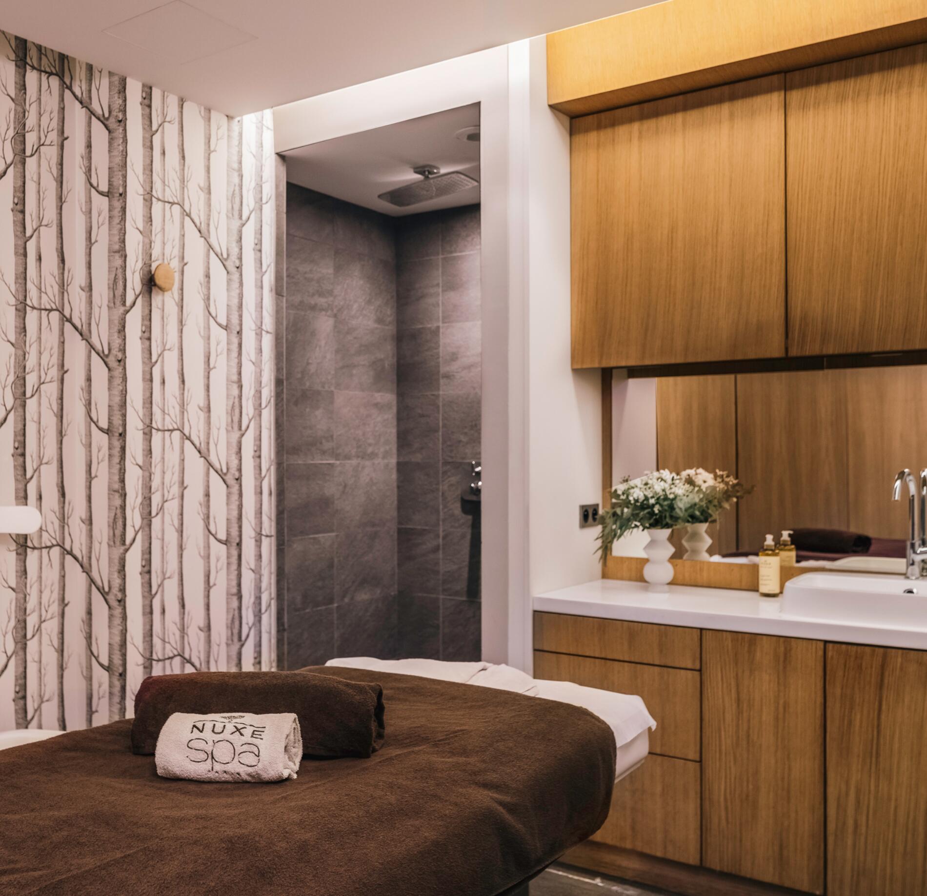 Cabine de soin élégante du spa NUXE à l’Hôtel Balthazar, avec table de massage soigneusement préparée, linge brun signature, espace lavabo en bois clair et décoration florale délicate, dans une atmosphère chaleureuse, calme et raffinée. 