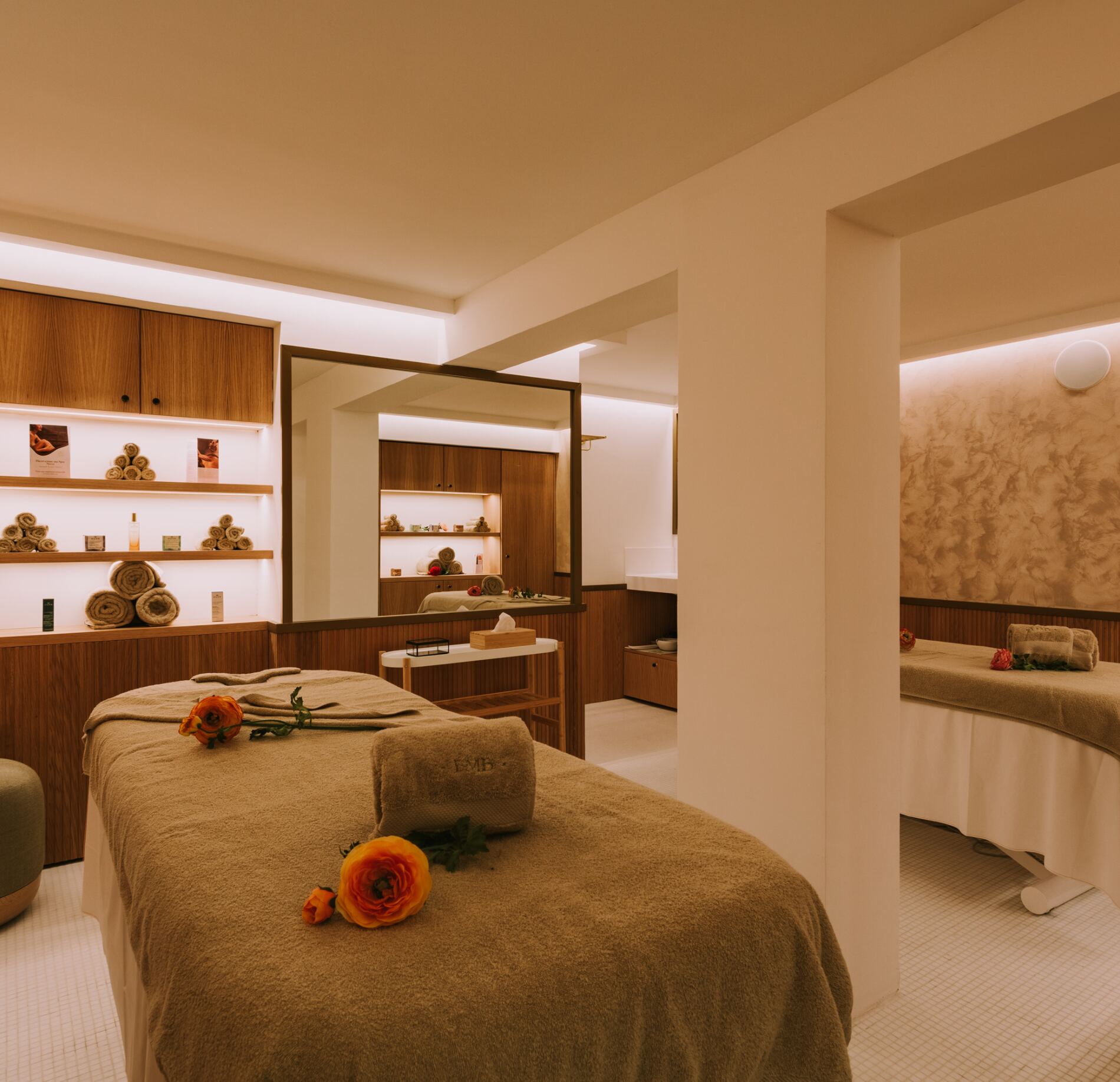 Cabine de soin lumineuse et élégante du spa NUXE, avec tables de massage soigneusement préparées, linge doux et fleurs délicates, entourées de mobilier en bois chaleureux et éclairage tamisé créant une atmosphère sereine et raffinée.