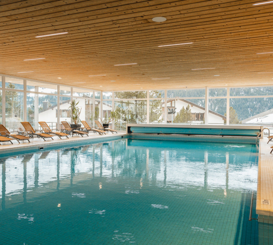 Grand bassin intérieur du spa NUXE à l’Hôtel Braunbär, bordé de transats confortables et baigné de lumière naturelle grâce aux larges fenêtres, avec plafond en bois chaleureux et vue sur les chalets et montagnes, offrant un cadre serein et haut de gamme propice à la détente.