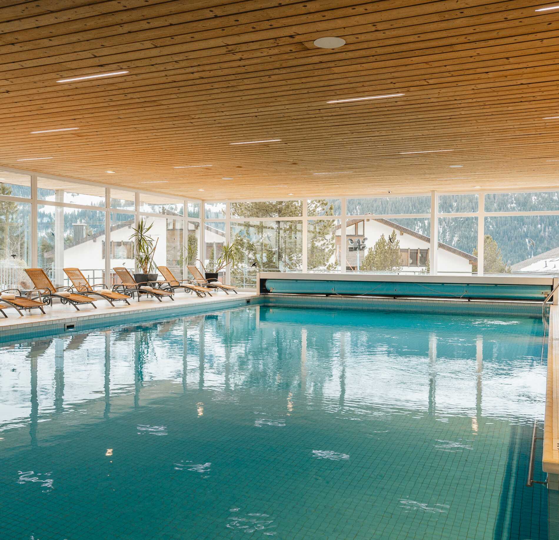 Piscine intérieure lumineuse du spa NUXE à l’Hôtel Braunbär, avec large bassin aux reflets turquoise, transats élégants alignés et grandes baies vitrées offrant une vue apaisante sur un paysage montagnard, créant une atmosphère chaleureuse et ressourçante. 