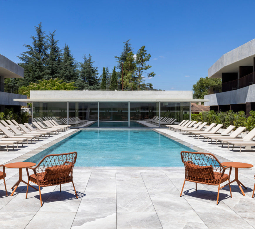 Un espace extérieur moderne du Spa La Villa Castellane, avec une grande piscine rectangulaire entourée de transats alignés symétriquement de chaque côté.
Au premier plan, deux fauteuils en métal aux tons orangés font face au bassin, tandis que des bâtiments contemporains et une végétation dense encadrent l’ensemble sous un ciel bleu clair.