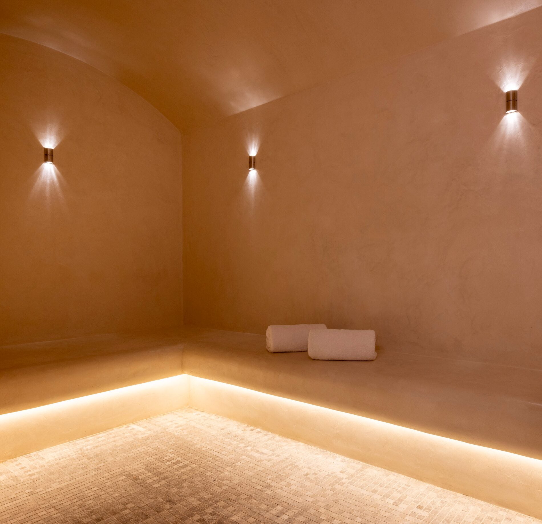 Un hammam minimaliste du Spa La Villa Castellane, avec des bancs en angle recouverts d’un enduit lisse aux teintes chaudes et deux serviettes blanches posées délicatement.
Un éclairage indirect sous les assises et des appliques murales diffusent une lumière douce, créant une atmosphère intime et enveloppante propice à la détente.