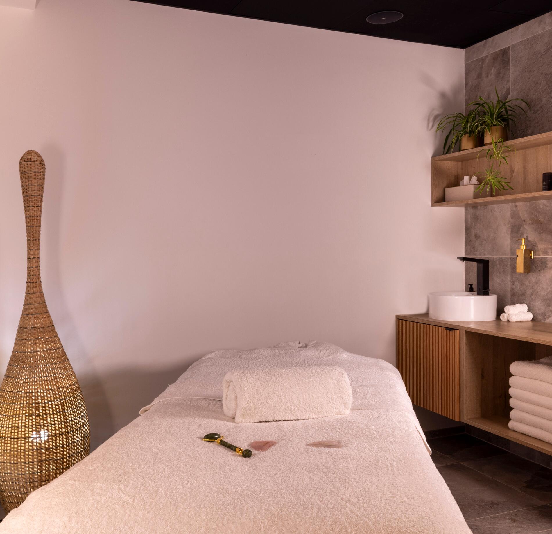 Une cabine de soin épurée du Spa La Villa Castellane, avec une table de massage recouverte de linge blanc et une serviette roulée placée au centre.
À côté, un meuble en bois avec vasque, étagères garnies de serviettes et plantes vertes, ainsi qu’un grand luminaire tressé, créent une ambiance douce et naturelle.