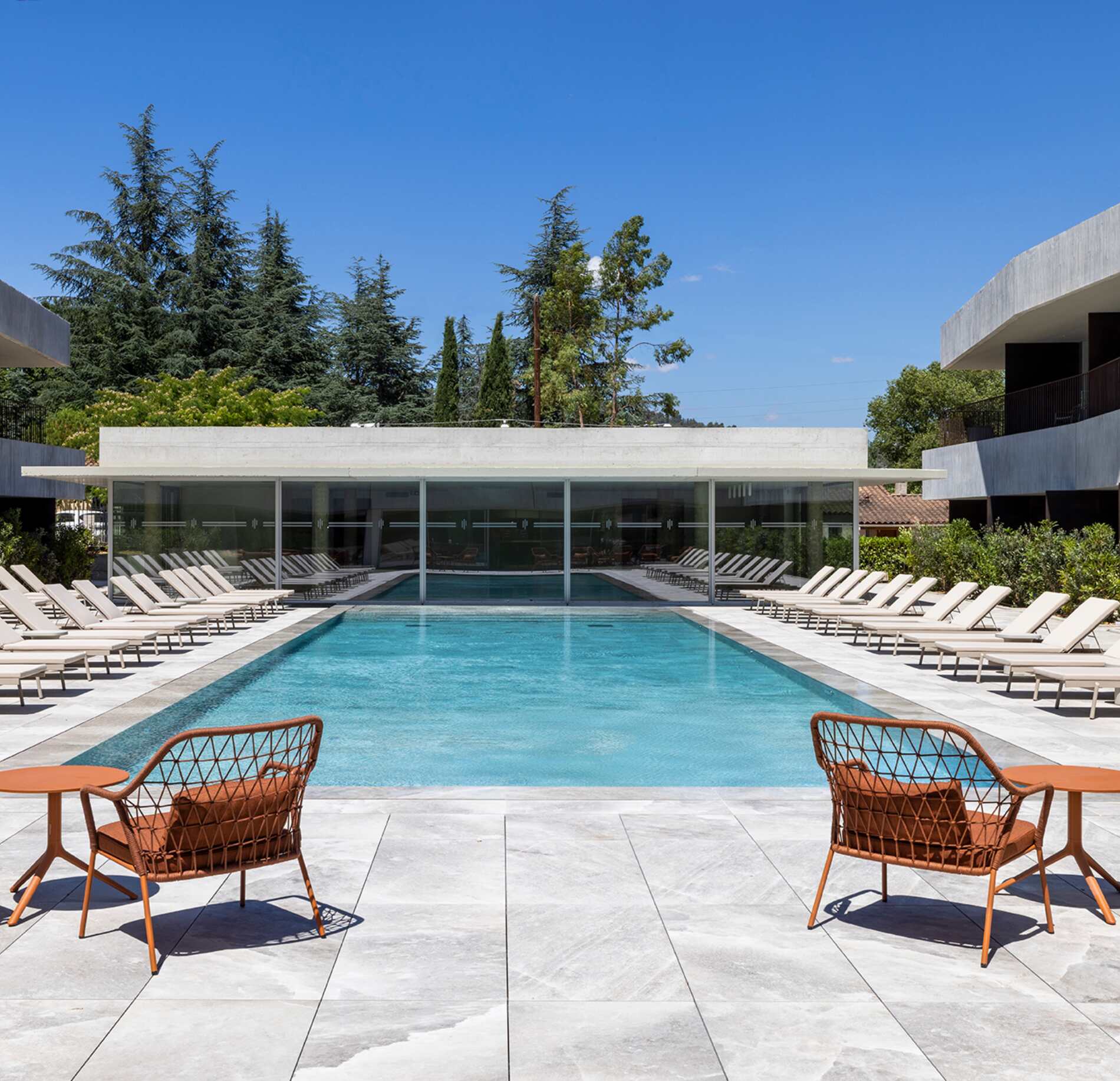 Un espace extérieur moderne du Spa La Villa Castellane, avec une grande piscine rectangulaire entourée de transats disposés symétriquement de chaque côté.
Au premier plan, deux fauteuils en métal aux tons orangés font face au bassin, tandis que des bâtiments contemporains et une végétation verdoyante encadrent l’ensemble sous un ciel bleu lumineux.
