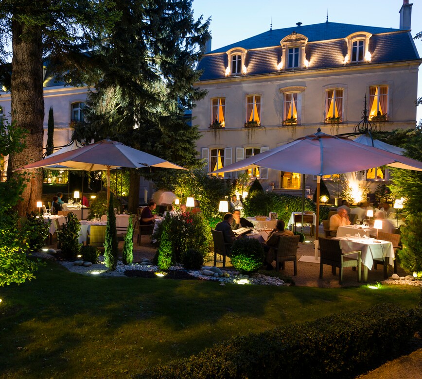 Une terrasse extérieure de l’Hostellerie Cèdre & Spa accueille plusieurs tables dressées sous de grands parasols, entourées de verdure et de lumière douce en soirée.
En arrière-plan, un élégant bâtiment aux fenêtres éclairées se dresse, donnant une atmosphère chaleureuse et raffinée à ce dîner en plein air