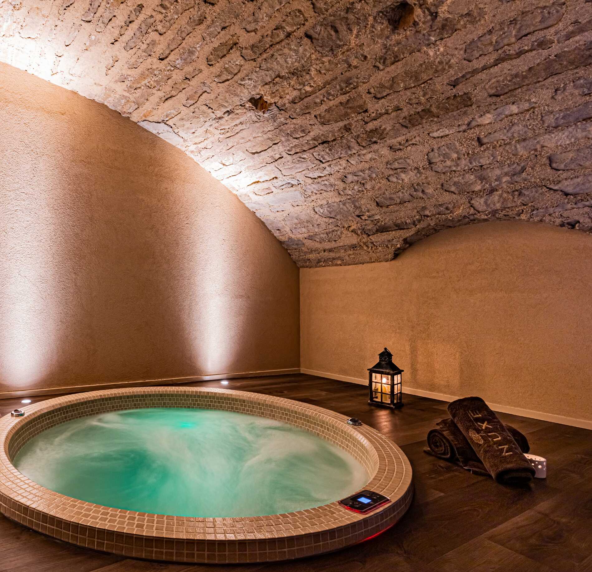 Un jacuzzi rond de l’Hostellerie Cèdre & Spa est encastré dans le sol d’une pièce voûtée en pierre, avec une eau turquoise en mouvement doux.
Autour, un sol en bois, une serviette roulée et une lanterne créent une ambiance intime et relaxante sous un éclairage chaud.