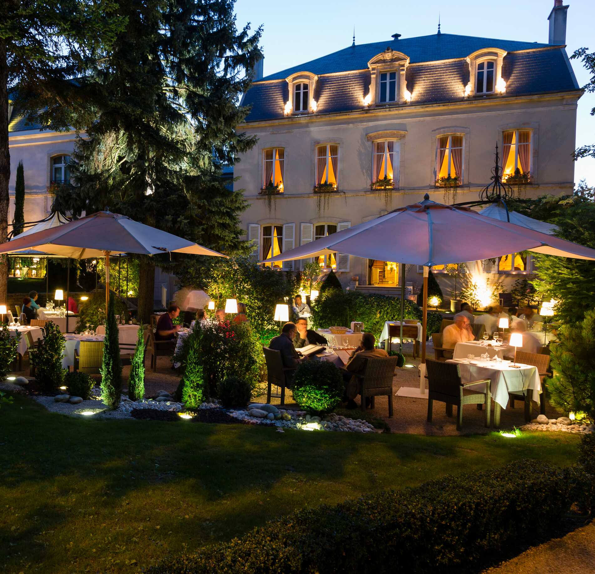 Une terrasse extérieure de l’Hostellerie Cèdre & Spa accueille plusieurs tables dressées sous de grands parasols, entourées de verdure et de lumière douce en soirée.
En arrière-plan, un élégant bâtiment aux fenêtres éclairées se dresse, donnant une atmosphère chaleureuse 