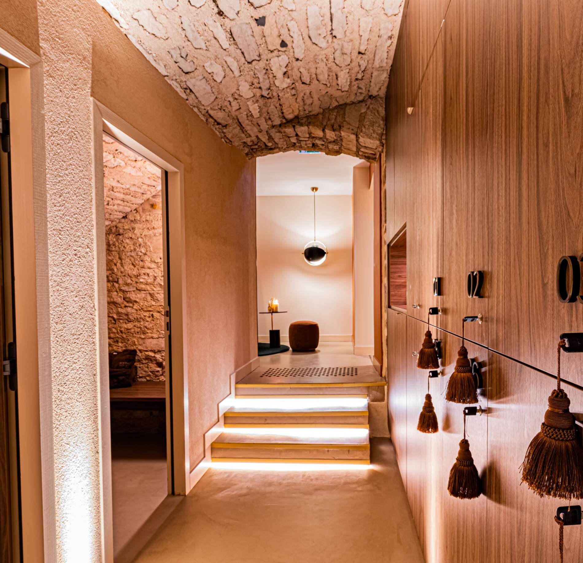 Un couloir intérieur de l’Hostellerie Cèdre & Spa présente des murs en bois et en pierre, avec un plafond voûté en briques apparentes et un éclairage doux au sol.
Sur la droite, des casiers numérotés avec des poignées en corde longent le mur, tandis qu’au fond quelques marches mènent vers un espace de détente épuré.