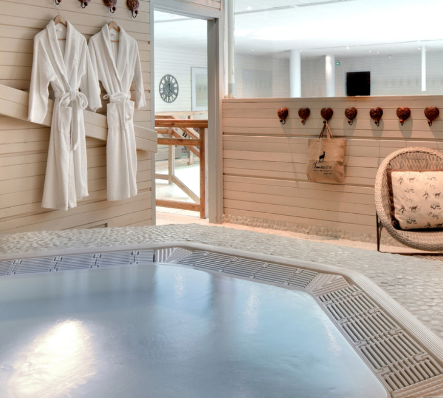 Espace détente lumineux du spa NUXE à l’Hôtel Le Chamois, avec jacuzzi aux eaux calmes au premier plan, peignoirs blancs soigneusement suspendus, crochets décoratifs et mobilier en bois clair, créant une atmosphère douce, chaleureuse et accueillante.