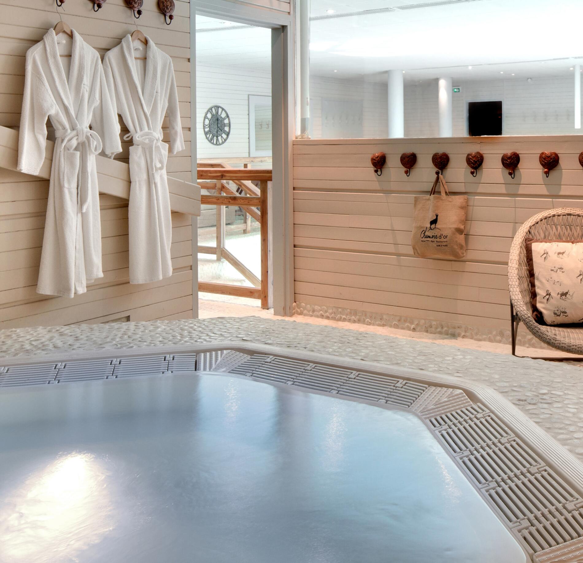Espace détente lumineux du spa NUXE à l’Hôtel Le Chamois, avec jacuzzi aux eaux apaisantes, peignoirs blancs suspendus et mobilier en bois clair, créant une atmosphère chaleureuse, élégante et cocooning. 