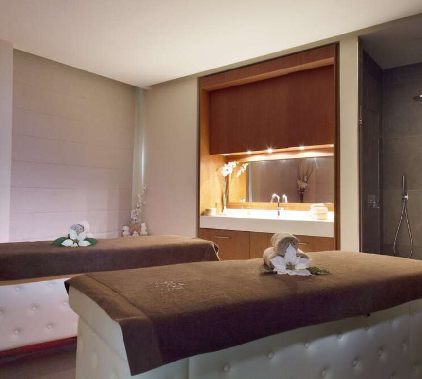 Une cabine de soin élégante du Relais Spa Val d'Europe, avec deux tables de massage recouvertes de linge brun et décorées de serviettes roulées et de fleurs.
À l’arrière, un espace avec meuble en bois, vasque et miroir éclairé diffuse une lumière douce, créant une atmosphère chaleureuse et raffinée.