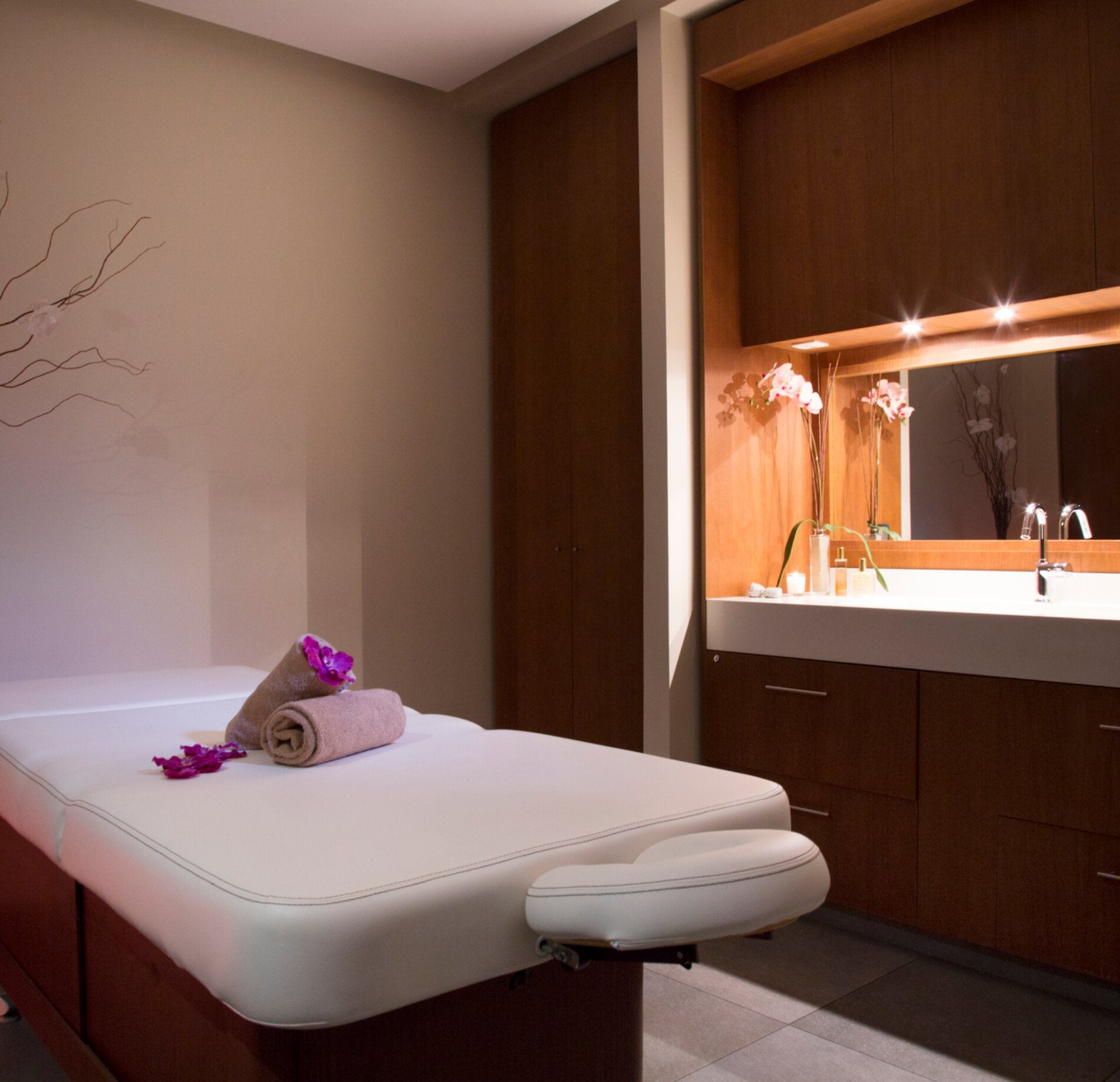 Une cabine de soin lumineuse du Relais Spa Val d'Europe, avec une table de massage recouverte de linge clair et décorée d’une serviette roulée accompagnée de fleurs.
À l’arrière, un meuble en bois avec vasque et miroir éclairé diffuse une lumière douce, créant une atmosphère élégante et apaisante.