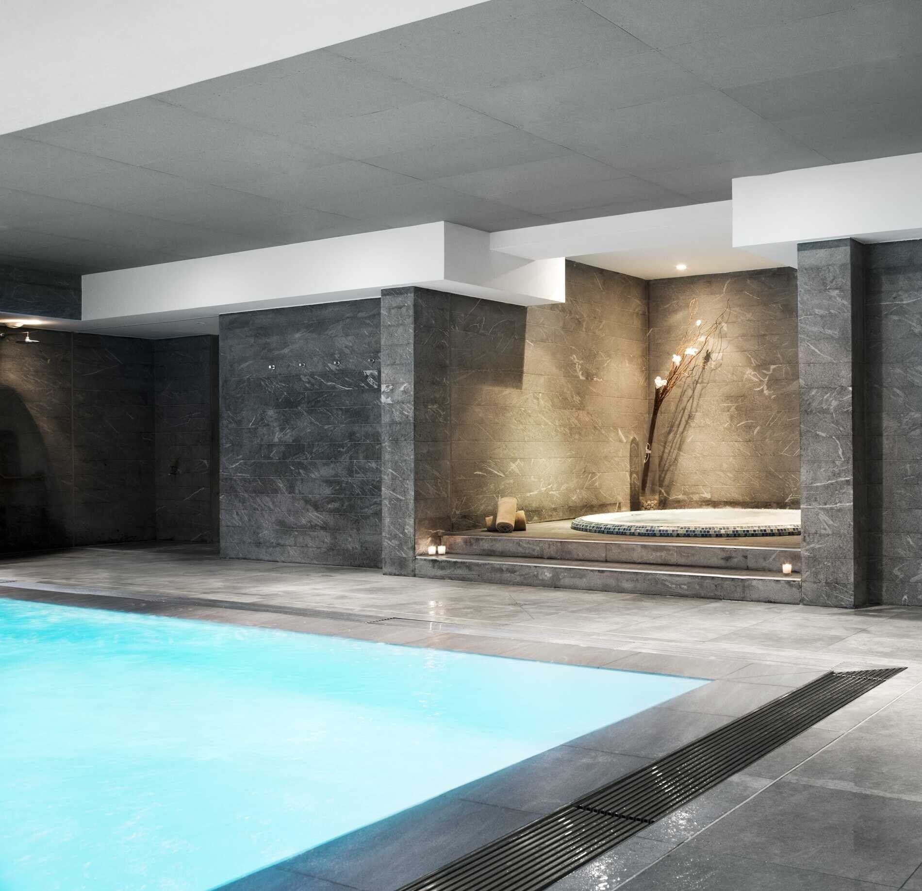 Un espace bien‑être contemporain du Relais Spa Val d'Europe, avec une piscine intérieure aux eaux bleu clair entourée de larges dalles en pierre grise.
À l’arrière, un jacuzzi légèrement surélevé et éclairé par des bougies s’intègre dans un décor minéral épuré, créant une ambiance calme et élégante.