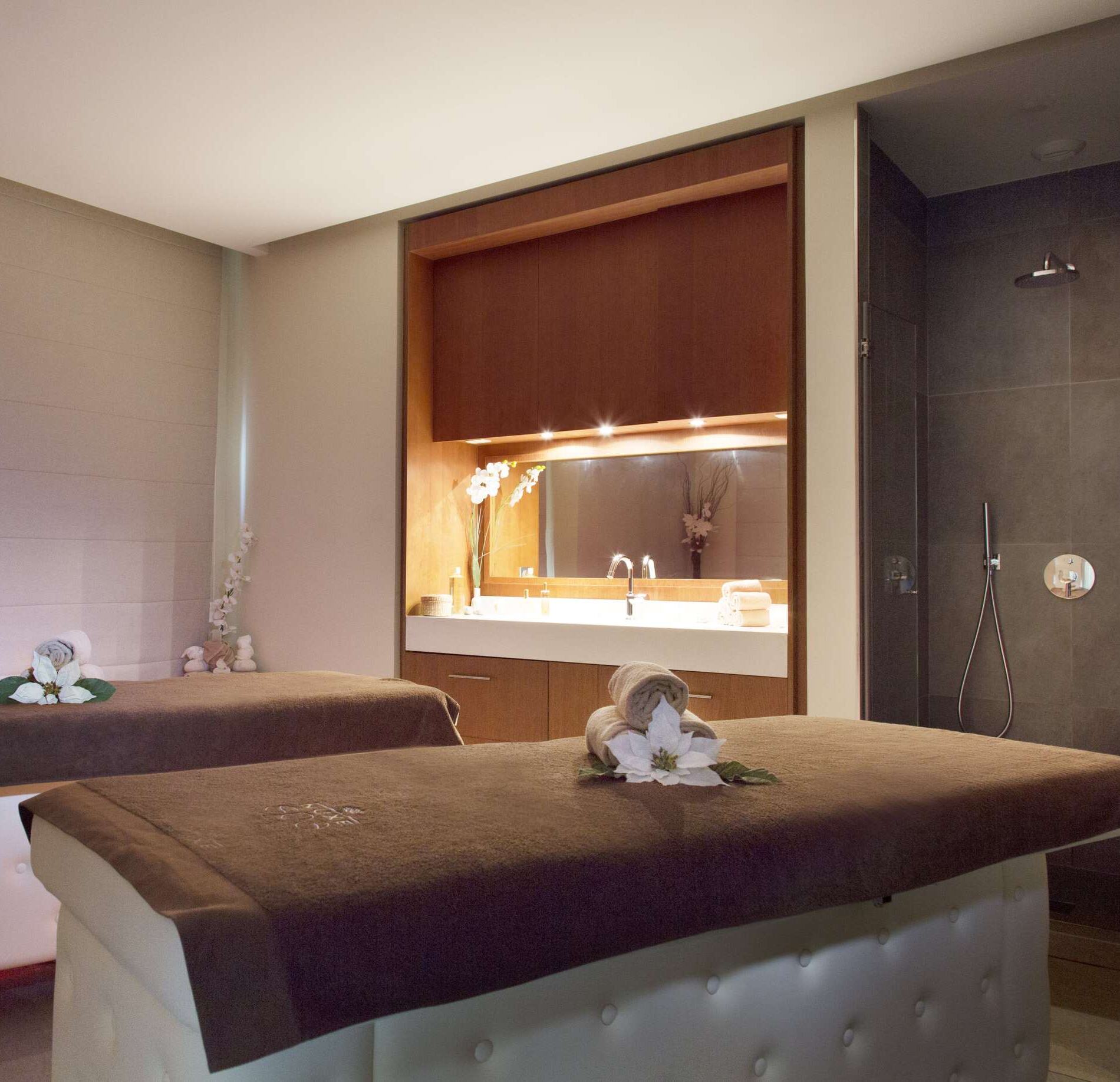 Une cabine de soin élégante du Relais Spa Val d'Europe, avec deux tables de massage recouvertes de linge brun et ornées de serviettes roulées accompagnées de fleurs.
À l’arrière, un espace avec meuble en bois, vasque et miroir éclairé diffuse une lumière douce, créant une ambiance chaleureuse et raffinée.